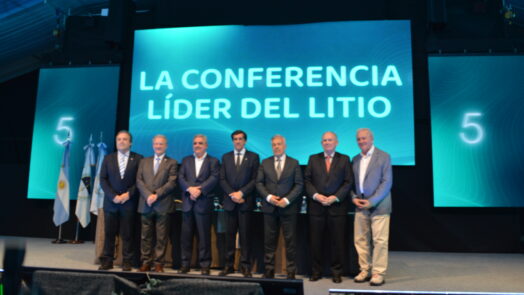 Siete participantes posan en el escenario de Litio en Sudamérica 2026, junto a banderas y una pantalla con la frase «La conferencia líder del litio».