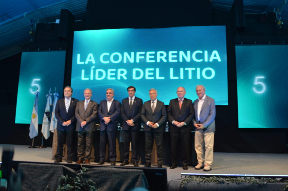 Siete participantes posan en el escenario de Litio en Sudamérica 2026, junto a banderas y una pantalla con la frase «La conferencia líder del litio».